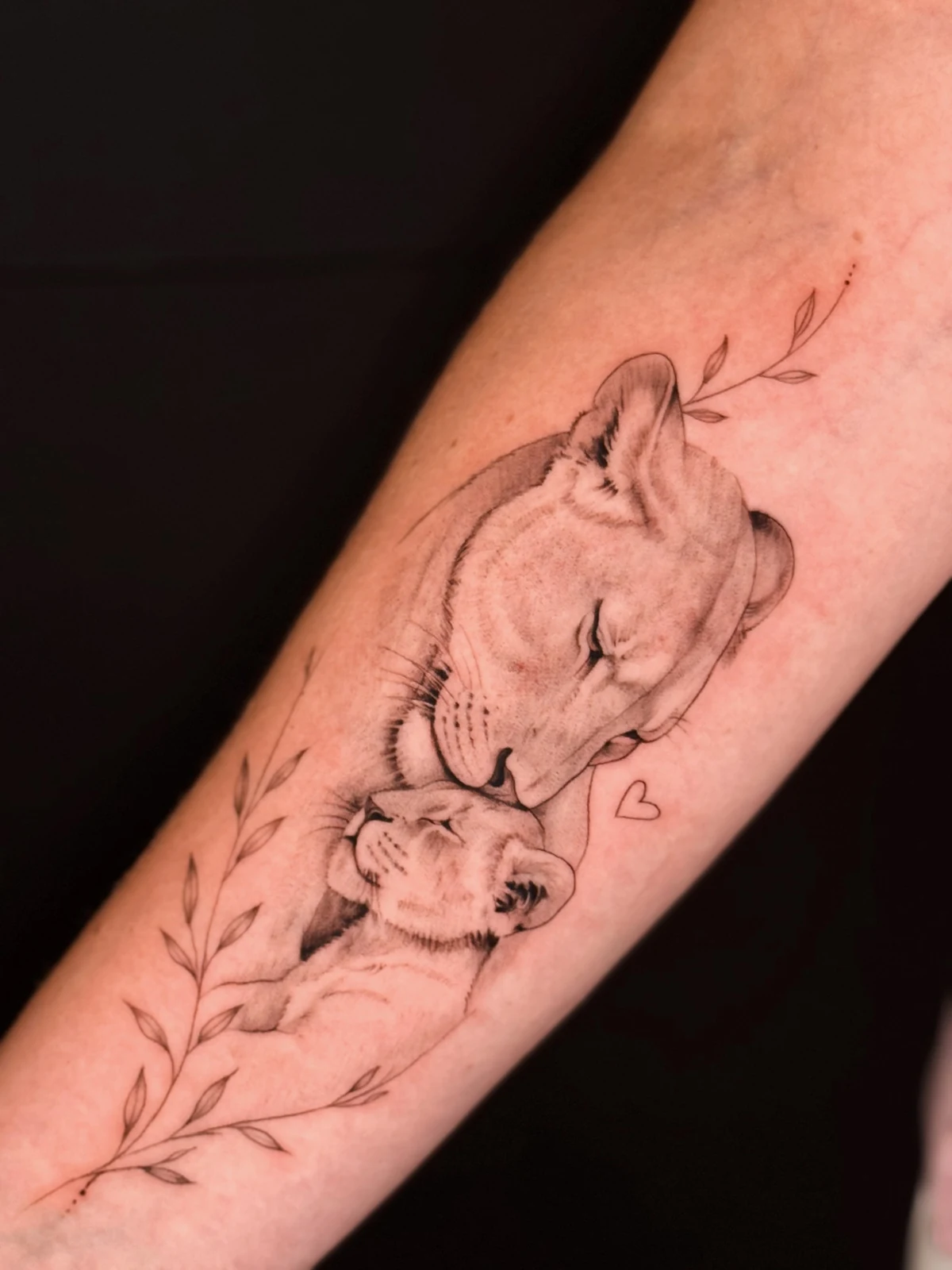 Black-and-Grey-Tattoo einer Löwin, die ihr Jungtier berührt, mit feinen Blätterranken am Unterarm – Arbeit von Merci, Skin City Tattoo.