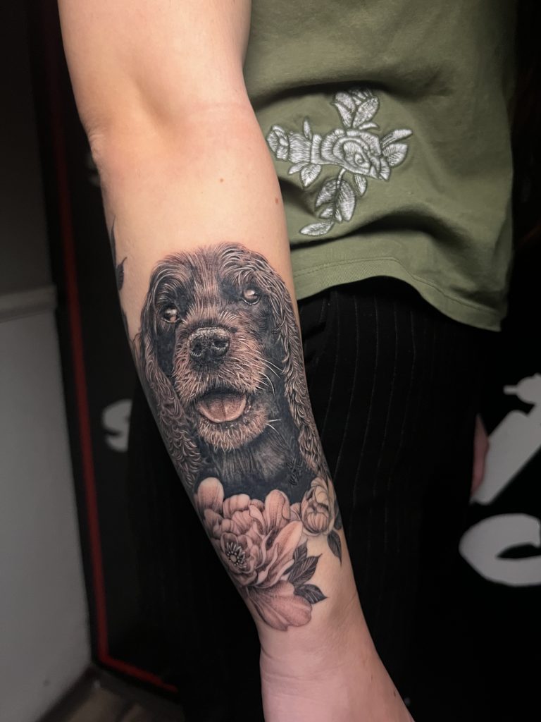 Realistisches Hunde-Tattoo mit Blüten in Schwarz und Grau am Unterarm
